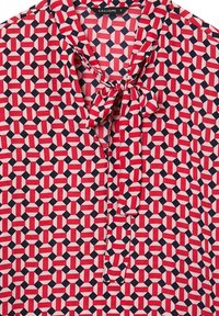 Blusa rossa e blu marina con motivo geometrico, collo a cravatta e tessuto morbido; presenta colori alternati in forme rettangolari.