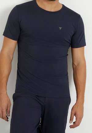 Uomo che indossa una t-shirt aderente a maniche corte blu navy con un piccolo logo triangolare Guess sul petto, abbinata a pantaloni blu navy.