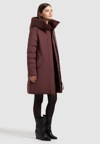 Bordový zimný kabát s vysokým golierom, vybavený bočnými vreckami a zoštíhleným pásom. Modelka má na sebe čierne klinové čižmy a pančuchy. Hladká textúra.