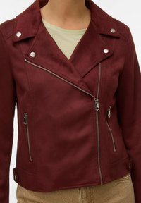 Femme portant une veste de motard bordeaux avec des fermetures éclair et des boutons-pression en argent, par-dessus un haut beige et un pantalon couleur sable.