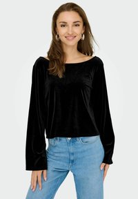 Top lungo in velluto nero con scollatura ampia e vestibilità rilassata, abbinato a jeans a vita alta blu chiaro.