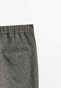 Gros plan d’un pantalon gris à motif chevrons montrant la ceinture et la poche passepoilée arrière sur fond blanc.