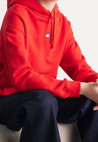 Czerwona bluza z kapturem, z przednią kieszenią kangurką, regulowanym sznurkiem i małym logo marki na klatce piersiowej. Połączona z ciemnymi dżinsami.
