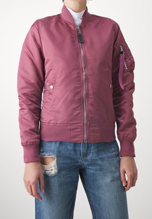 Personne portant une veste bomber violette zippée avec des poignets côtelés et une poche zippée sur la manche, associée à un jean bleu déchiré.