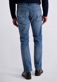 Rückansicht einer Person, die blaue gerade geschnittene Jeans mit Gesäßtaschen und dunkelbraunen Schuhen auf einem hellen Hintergrund trägt.