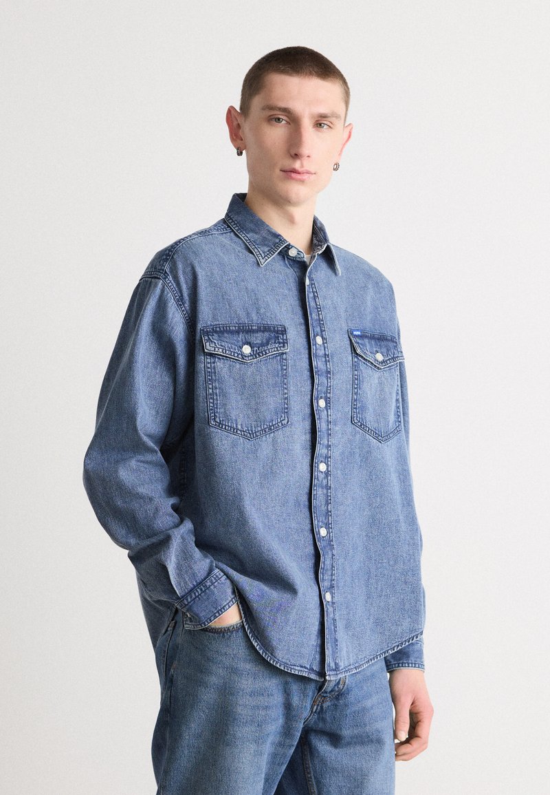 Denim overhemd met lange mouwen, knoopsluiting aan de voorkant, twee borstzakken, lichtblauwe kleur, zware stof, en contrasterende stikseldetails.