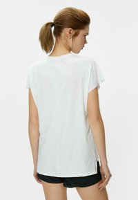 Top blanco de manga corta con un ajuste relajado, textura suave y aberturas laterales, combinado con pantalones cortos negros. Vista trasera que destaca un diseño sencillo.