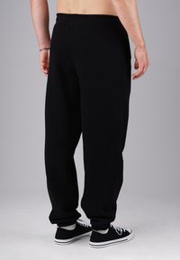 Male wearing black sweatpants and black sneakers with white soles and laces, standing against a plain gray background.