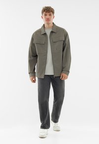 Jasnoszara kurtka z rozłożonym kołnierzem i dwoma przednimi kieszeniami, noszona na jasnej koszuli i ciemnych dżinsach, w połączeniu z białymi trampkami.