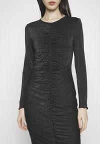 Femme portant une robe noire cintrée à manches longues avec des détails froncés au centre et un tissu subtilement scintillant.