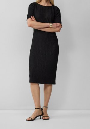 Femme portant une robe noire texturée longueur genou, des sandales noires à talons et un bracelet en or, debout avec les bras croisés sur un fond gris.