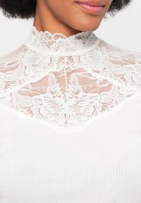 Top blanco con un alto cuello de encaje con patrones florales y un cuerpo acanalado. El encaje es intrincado y semitranslúcido, creando un contraste texturizado.