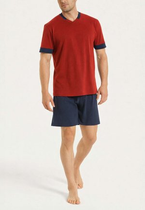 Uomo che indossa una maglietta rossa a maniche corte con polsini e collo blu navy, abbinata a pantaloncini blu navy, che cammina a piedi nudi su uno sfondo chiaro.