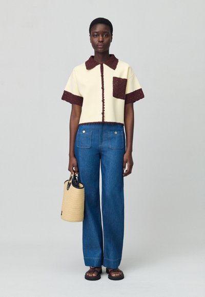 Suéter de manga corta color crema con ribete burdeos y bolsillo, combinado con pantalones de mezclilla anchos de color azul y un bolso de palma tejido. Sandalias negras completan el look.