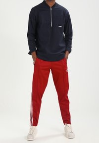 Tmavě modrý top s dlouhým rukávem, zipovým límcem a kapsou, doplněný červenými tepláky s bílými pruhy a logem, zakončený bílými teniskami.