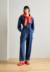 Macacão de ganga com mangas longas, cintura amarrada e bolsos de cargo; combinado com sandálias vermelhas de salto grosso e um suéter vermelho correspondente amarrado no pescoço.