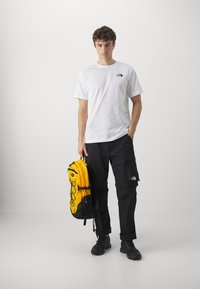 Hvit t-skjorte med korte ermer, svarte cargo-bukser, holder en lys gul ryggsekk med svarte detaljer og blomsterdesign. Svarte sko.