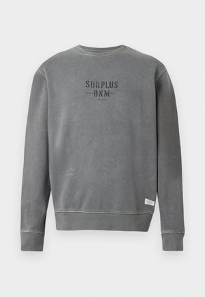 Harmaa pitkähihaisuus crewneck-huppari, jossa rintapielessä mustalla printattuna "SURPLUS DNM" -teksti ja pieni valkoinen etiketti helman lähellä.