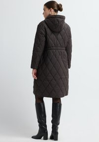 Donkerbruine gequiltte jas met een capuchon, een getailleerde taille en zijzakken. Afgewerkt met een gladde textuur en lange mouwen. Gedragen met kniehoge zwarte laarzen.