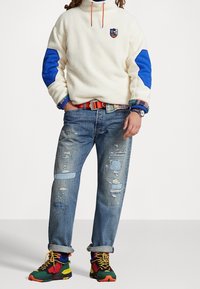 Fleecepullover i off-white med blå armbågsfärger, blå dragsko och logotyp. Bärs med slitna blå jeans och färgglada sneakers.