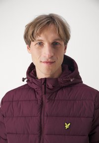 Bordo puffer jakna s kapuljačom, s horizontalnom kviltanom teksturom i malim žutim ptičjim logotipom s lijeve strane prsnog dijela.
