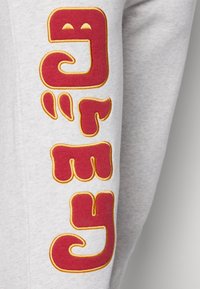 Grå sweatshirt med stora, broderade röda bokstäver och gula accenter på vänster ärm. Texten har en lekfull, rundad design.