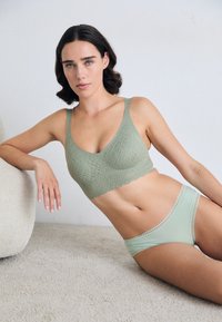 Žena leži na koberci, oblečená v svetlozelenej vzorovanej bralette a zodpovedajúcom spodnom prádle s čipkovým lemovaním, s krátkymi tmavými vlasmi a neutrálnym výrazom tváre.