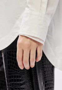 Λευκό μανίκι πουκάμισου με λεία υφή και κουμπωτό μανσέτα, σε αντίθεση με ένα χέρι που ξεκουράζεται δίπλα από υφασμάτινο παντελόνι μαύρου χρώματος με σχέδιο κροκοδείλου.