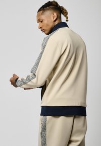 Veste de sport beige avec col marine, présentant des manches à motifs et un tissu texturé. La coupe est décontractée avec des détails de couture visibles.