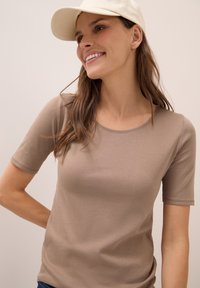 Kratší béžové tričko s krátkým rukávem a kulatým výstřihem, vyrobené z měkké látky, přiléhavého střihu a decentním švem. Na sobě má bílé čepice.