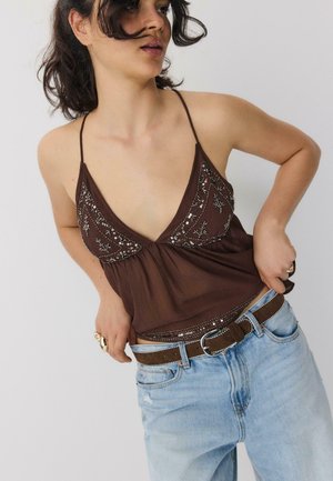 Donna che indossa un top marrone con paillettes e jeans azzurri che si sistema il top con una mano sulla vita, su uno sfondo semplice.