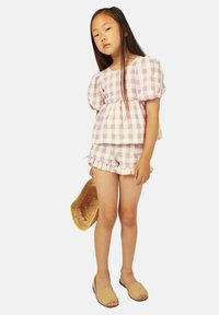Conjunto de dos piezas a cuadros en tonos morado claro y blanco, que incluye una blusa con mangas abullonadas y pantalones cortos con volantes, combinados con sandalias tipo slip-on en color beige.