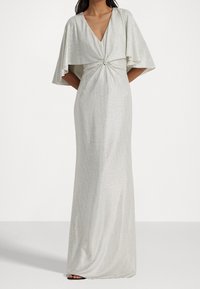 Kvinna klädd i en golvsläpande, silver metallic klänning med en djup V-neck och breda, cape-liknande ärmar, stående mot en enkel bakgrund.