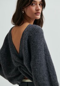 Žena s tmavými vlasy v texturovaném tmavošedém svetru s hlubokým véčkovým výstřihem na zádech a zkrouceným detailem, stojící na pozadí s jemným světlým tónem.