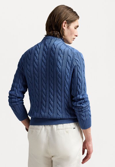 Blå kabelstrikket sweater med rund hals, ribbede ærmer og nederkant. Båret over cremede bukser, der viser struktureret stof og designdetaljer.
