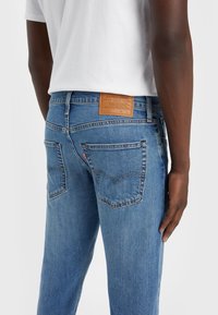 Jeans en denim bleu à coupe droite, avec un patch en cuir à l'arrière et deux poches arrière avec des détails cousus. Tissu délavé et texturé.