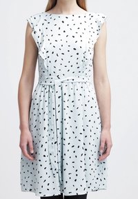 Femme portant une robe blanche sans manches avec des pois noirs irréguliers, des détails en volants aux épaules et une ceinture nouée à la taille.