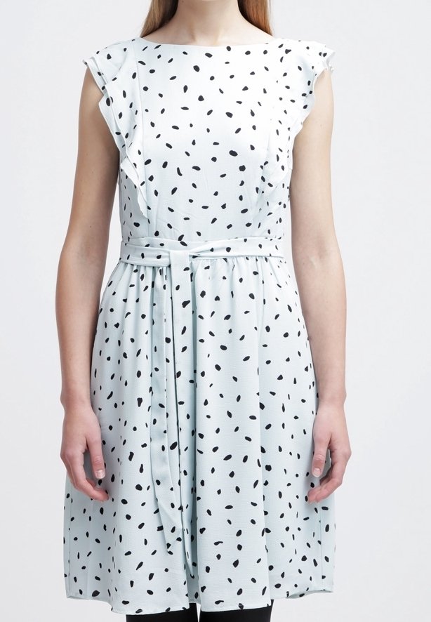 Femme portant une robe blanche sans manches avec des pois noirs irréguliers, des détails en volants aux épaules et une ceinture nouée à la taille.