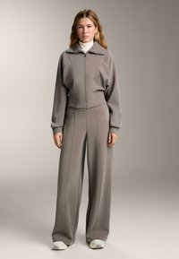 Szürke sportos szett, zipzáras crop dzsekivel, amelynek gallérja van, valamint laza nadrágokkal, mindkettő puha anyagból készült, kontrasztos varrási részletekkel.