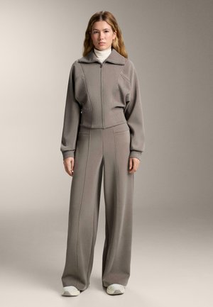 Šedý športový set pozostávajúci z kabátika s krátkym zipsom a golierom a nohavíc s širokým strihom, obidva z mäkkej tkaniny s kontrastným prešívaním.