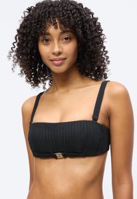 Γυναίκα με σγουρά μαλλιά που φοράει μαύρο ριμπ μπούστο μαγιό με λεπτές τιράντες και χρυσό κούμπωμα στο κέντρο, σε απλό λευκό φόντο.