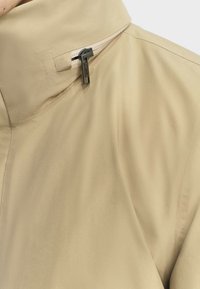 Beige Jacke mit hohem Kragen, glatter Textur und einem auffälligen Reißverschlussdetail am Hals. Klare Linien und minimalistisches Design.