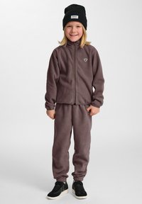 Fleece-träningsdräkt i dämpad lila, med en zip-jacka och elastiska muddAr, tillsammans med svarta sneakers och en svart mössa.