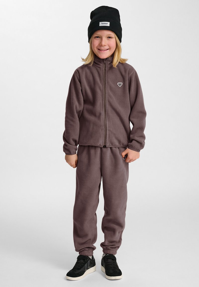 Fleece-träningsdräkt i dämpad lila, med en zip-jacka och elastiska muddAr, tillsammans med svarta sneakers och en svart mössa.