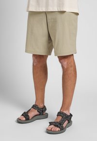 Svarte sandaler med mønstrede remmer og grå såler, kombinert med lysebeige kne-lange shorts, som avdekker teksturen på den nedre delen av benet.