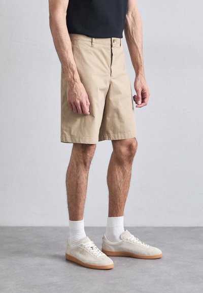 Pantaloni scurți din bumbac bej, cu croială dreaptă, având un fermoar frontal și închidere cu nasture. Purtați cu șosete albe și adidași gri deschis.