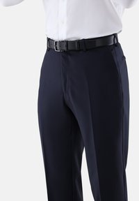 Mann trägt marineblaue Stoffhose mit schwarzem Gürtel und weißem, in die Hose gestecktem Hemd, von der Taille bis zur Mitte des Oberschenkels zu sehen.