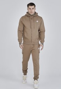 Ruda zip-up džinsinė striukė su priekyje esančia kišene, derinama su atitinkančiais cargo stiliaus džinsais, turinčiais elastingas rankoves ir logotipo akcentus, avimi baltais sportbačiais.