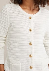 Vit stickad kofta med horisontella ränder, prydd med fem knappar i guldton och två framfickor. Texturerad tyg och rundad halsringning.