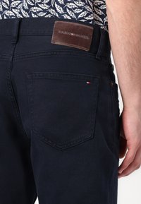 Jeans in denim blu scuro con una toppa in pelle etichettata "TOMMY HILFIGER." Presentano una tasca posteriore con un piccolo accento rosso e bianco.